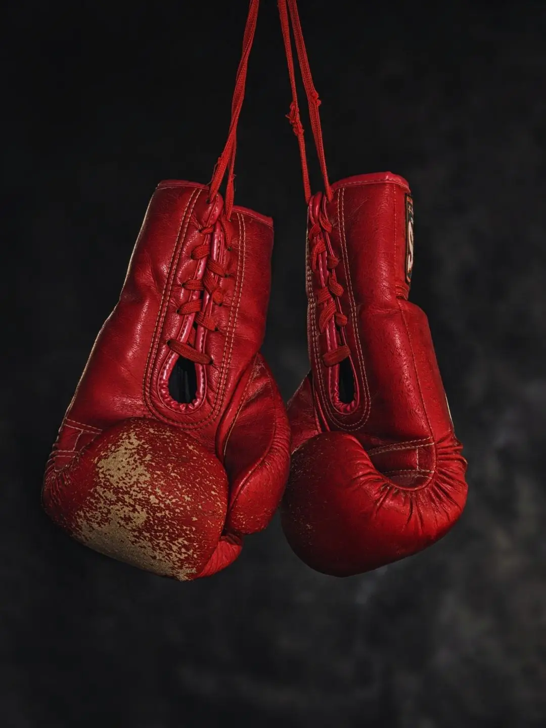 A pair of worn red leather boxing gloves hanging by their laces against a dark background.